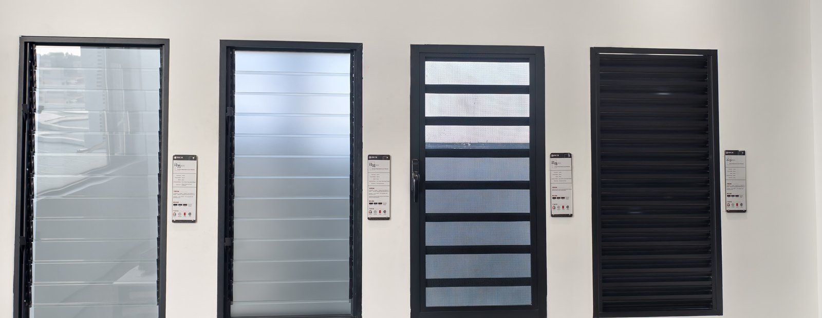 Four black-framed window systems mounted side by side on the showroom sample wall — a screen, a roller shutter, a louvre with a screen and a louvre blind