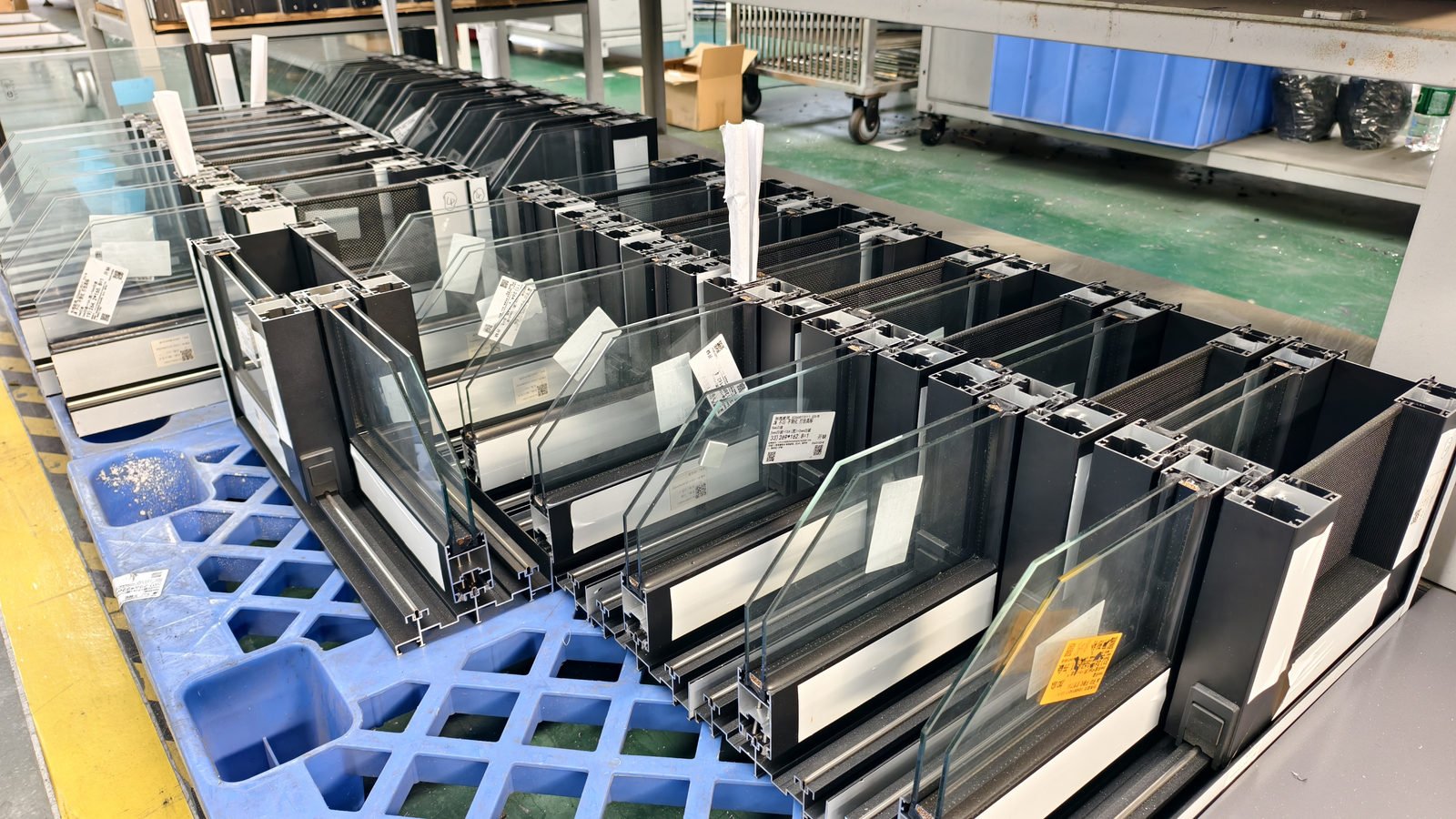 Black window frames stacked on blue pallets on the factory floor
